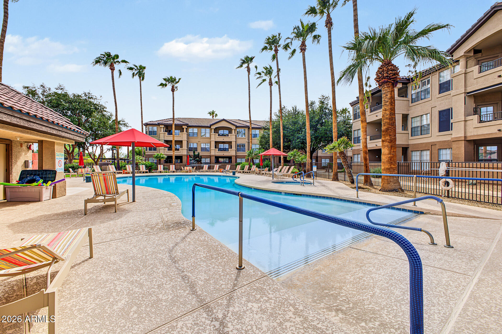 Community swimming pool with apartment buildings, palm trees, and red shade umbrellas.