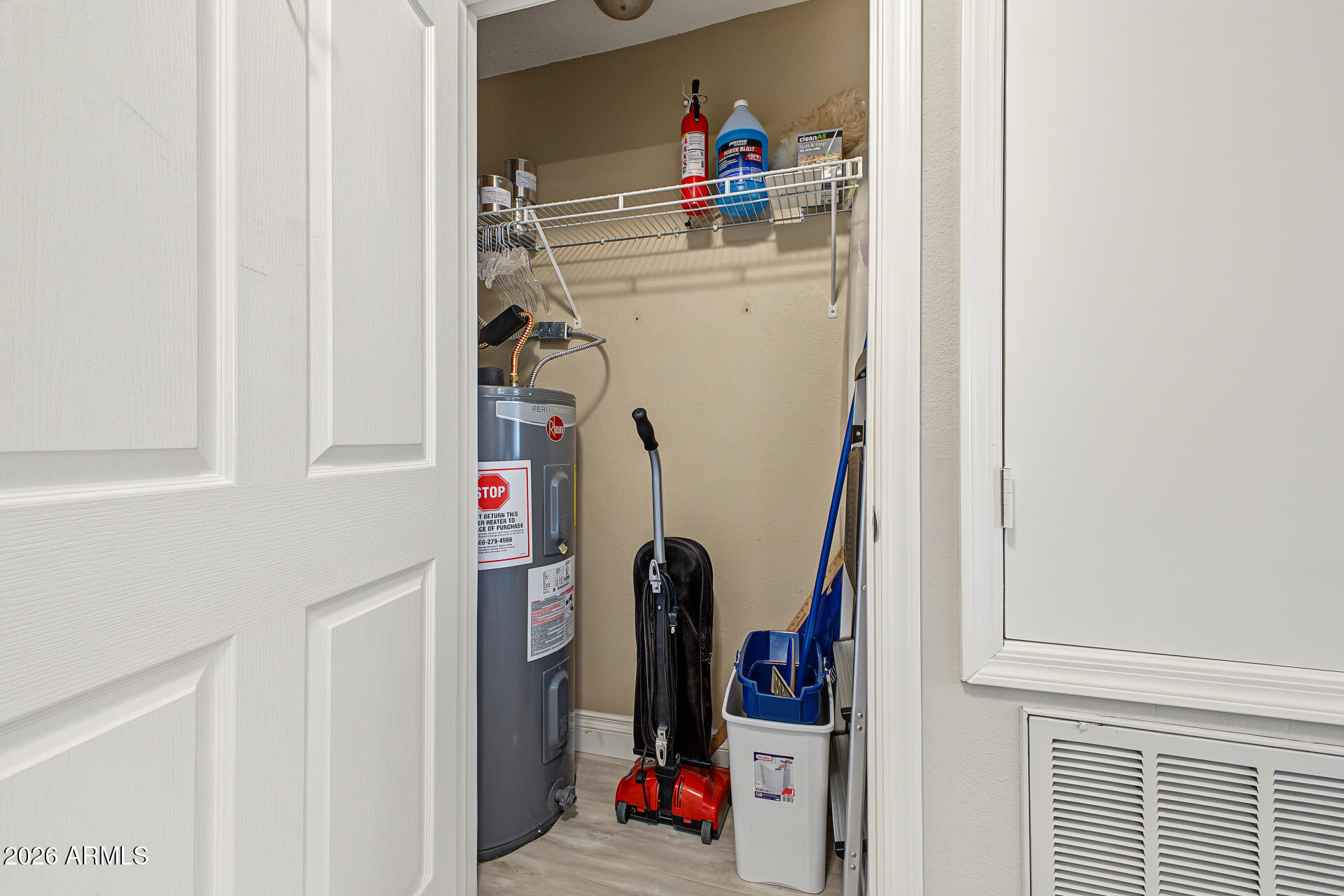 Utility closet with a water heater, cleaning supplies, vacuum, and overhead wire shelf.