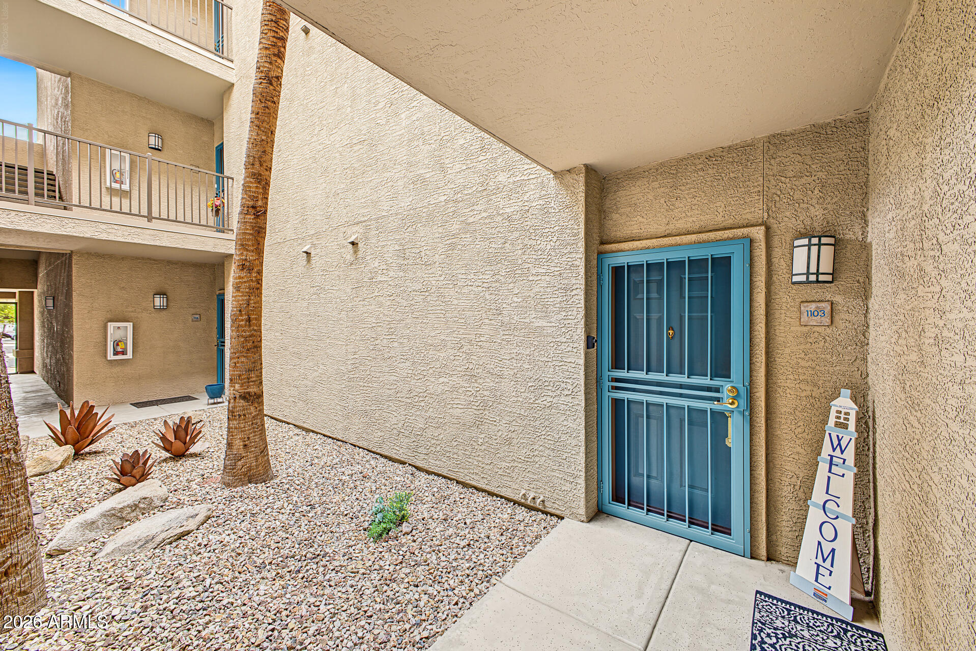 Apartment entrance with a bright blue security door, gravel landscaping, and a welcome sign.