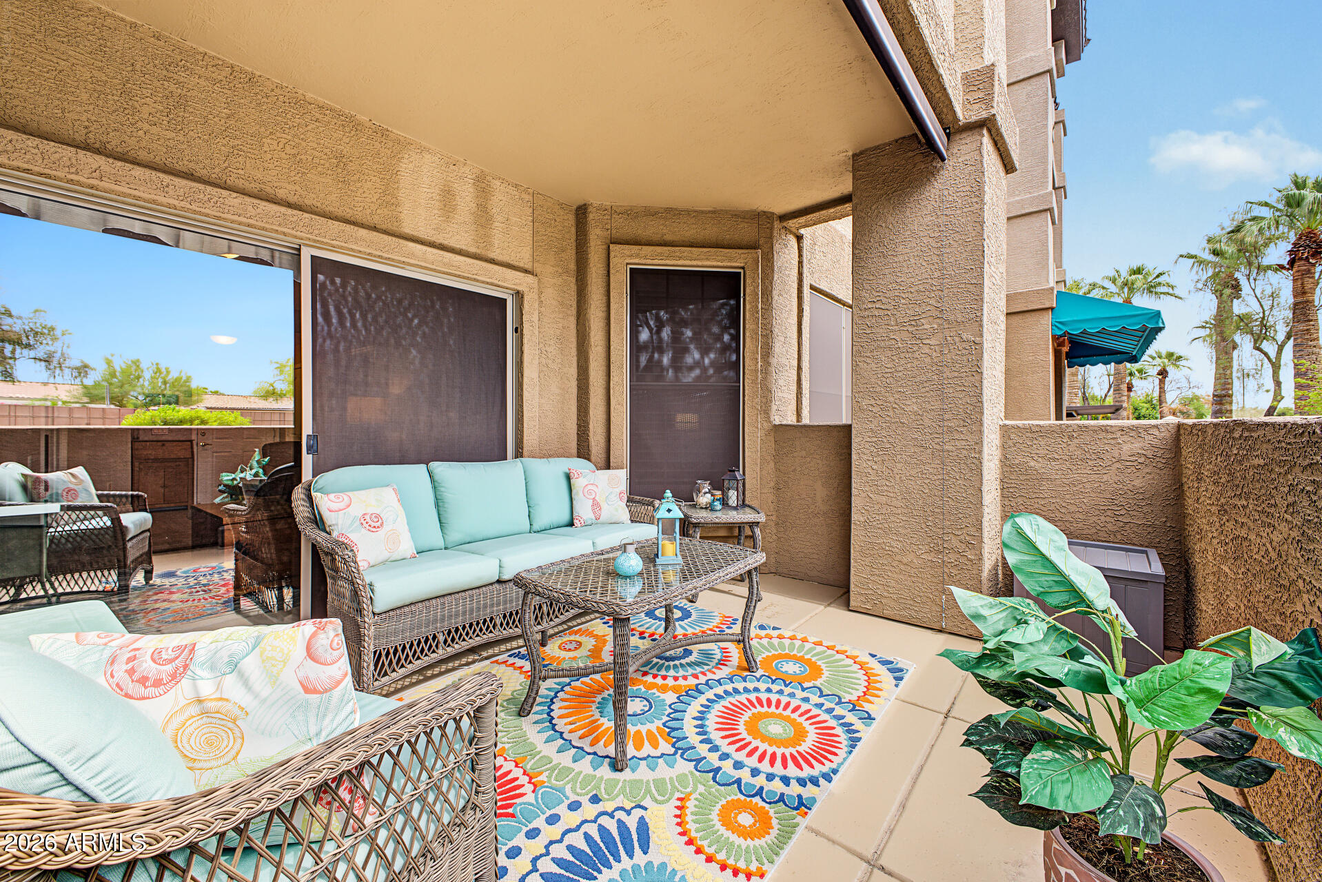 Covered patio with turquoise wicker seating, a colorful floral rug, and potted plants.