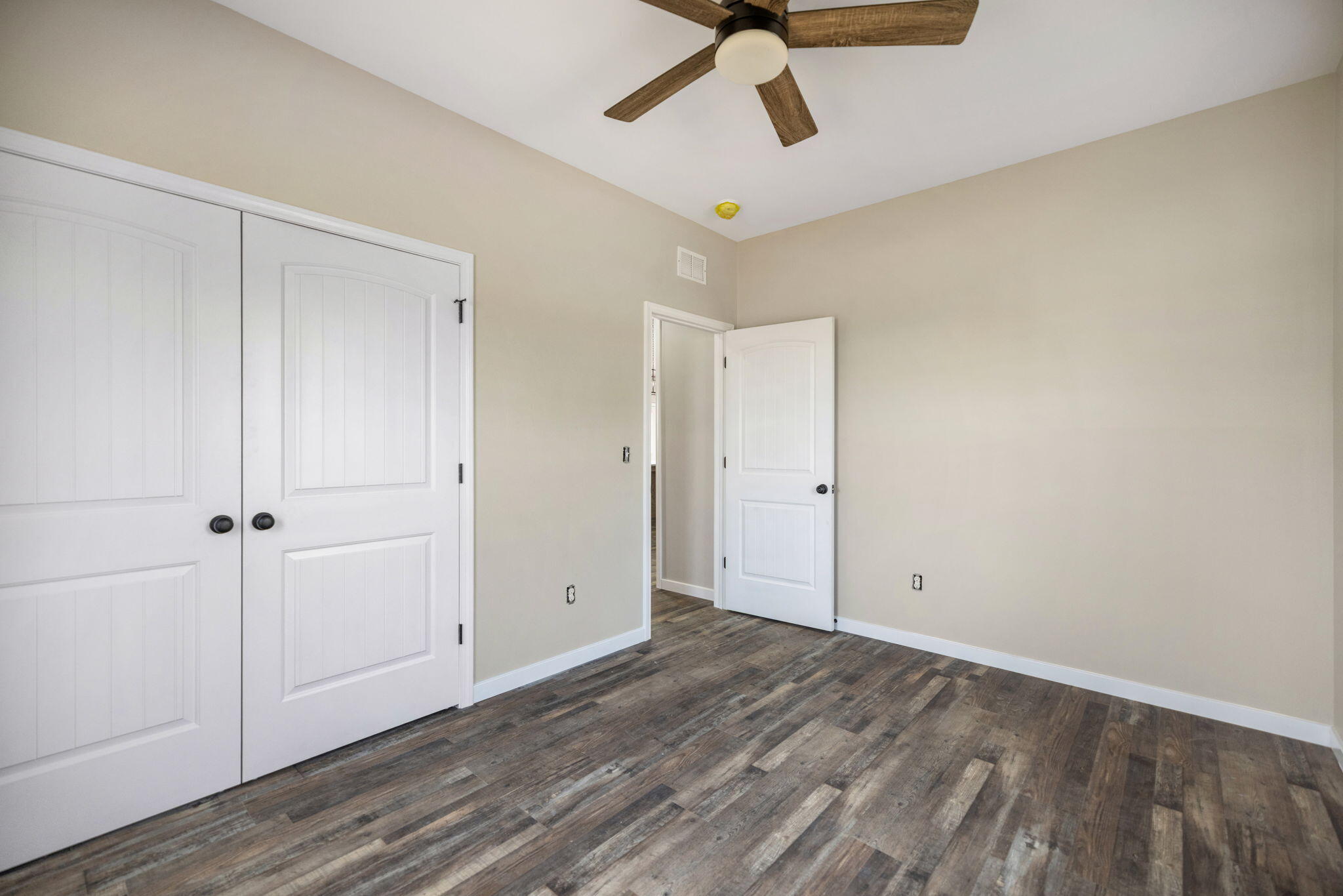 Empty bedroom with white paneled closet doors, wood-look flooring, and a ceiling fan.
