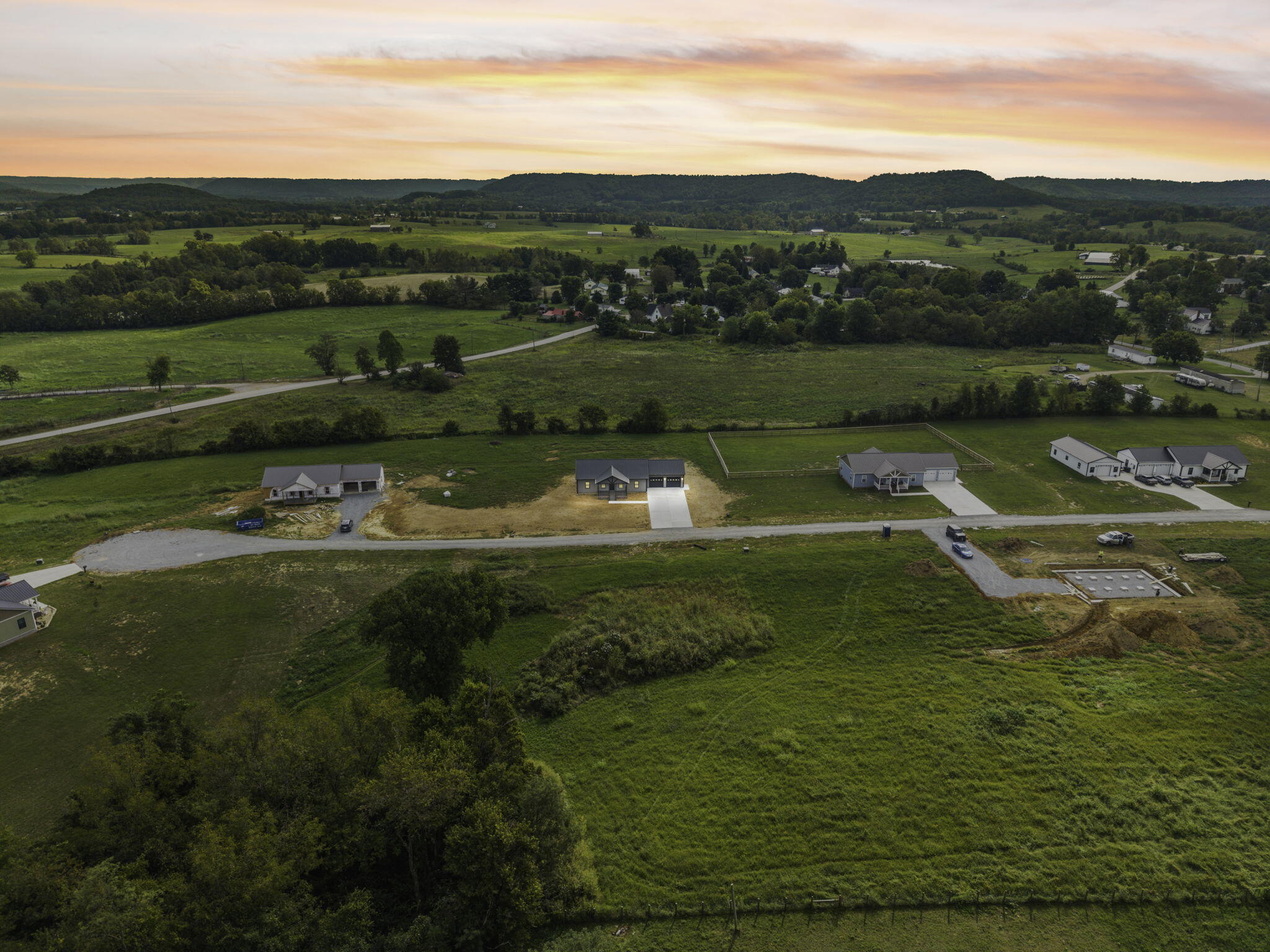 Aerial view of homes and building sites along a rural road, surrounded by green fields and wooded hills.