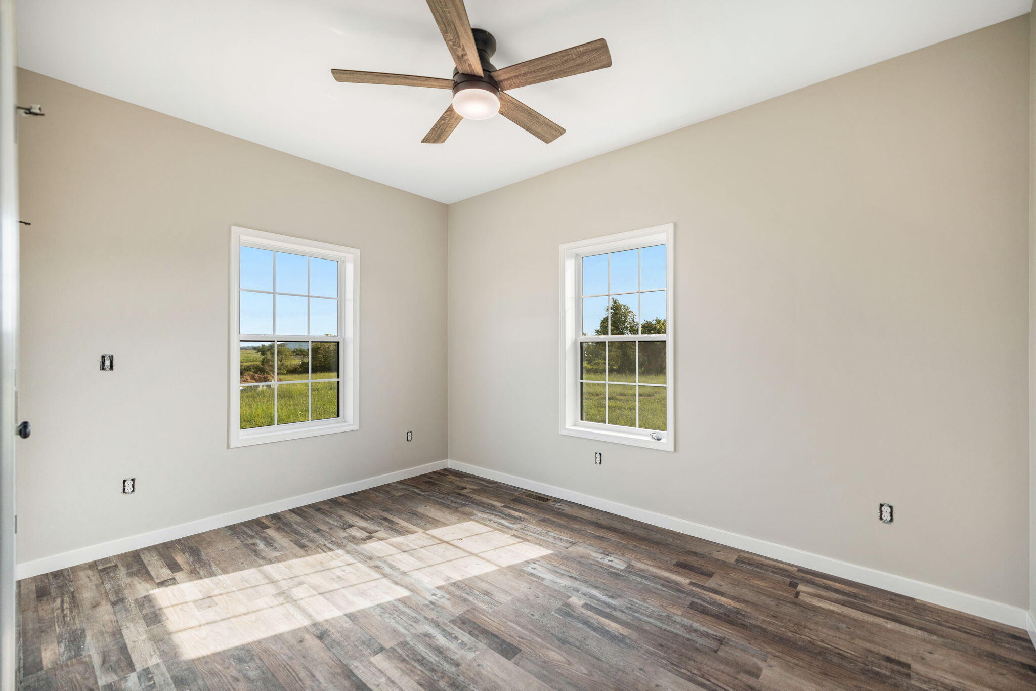 Vacant room with two windows, a wood-blade ceiling fan, and sunlight across plank flooring.