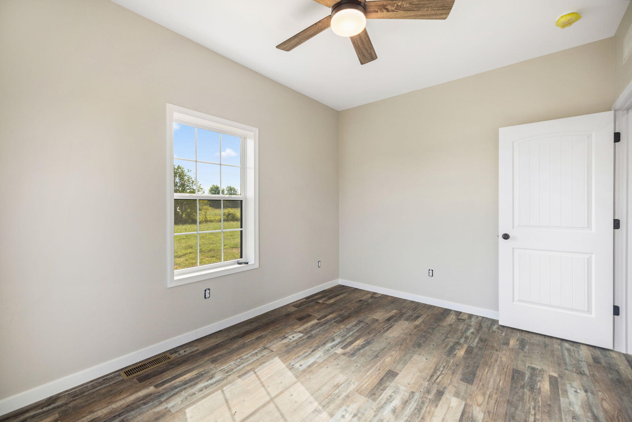 Empty room with wood-look flooring, a window, open white door, and ceiling fan.