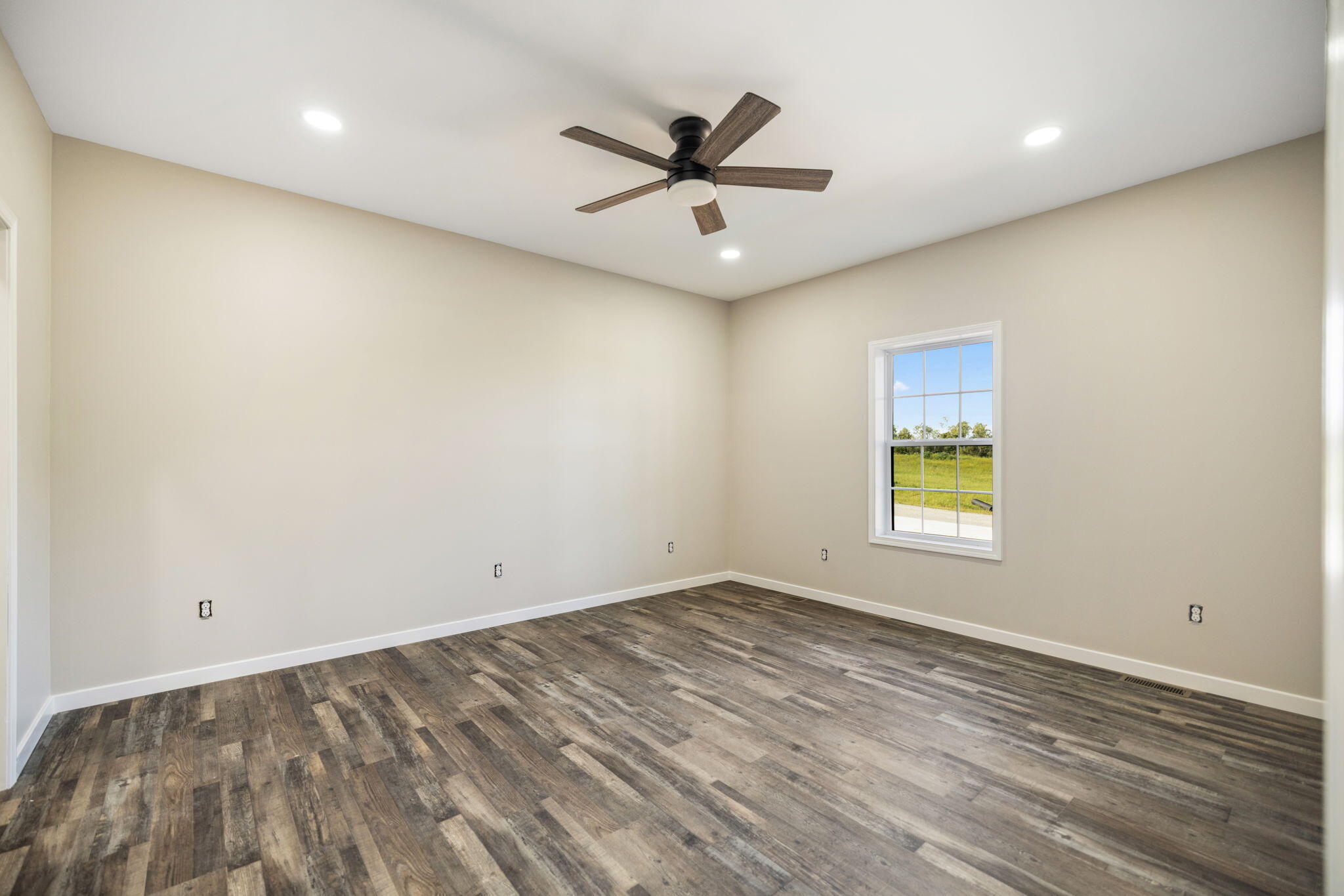 Empty room with wood-look flooring, a dark ceiling fan, and a window overlooking grass.