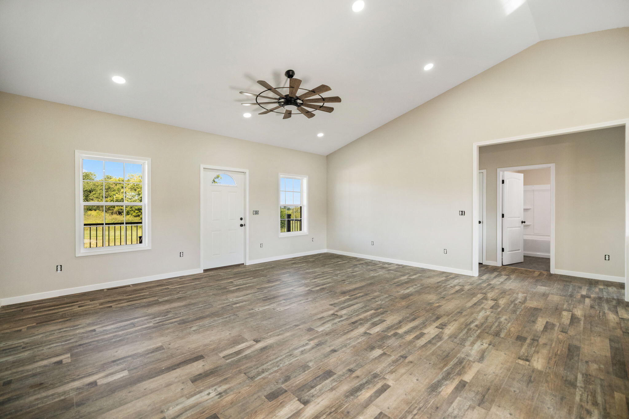 Vacant living room with wood-look floors, vaulted ceiling, ceiling fan, and two windows.
