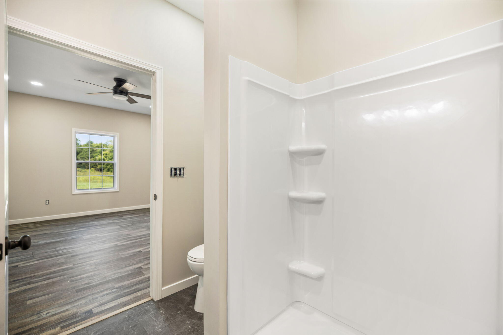 Bathroom shower with built-in shelves beside a doorway to an empty room with gray plank flooring.