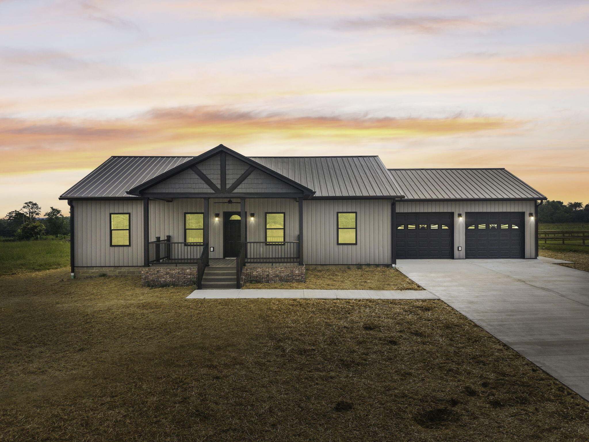 Single-story gray home with a covered porch, dark metal roof, and two garage doors at sunset.