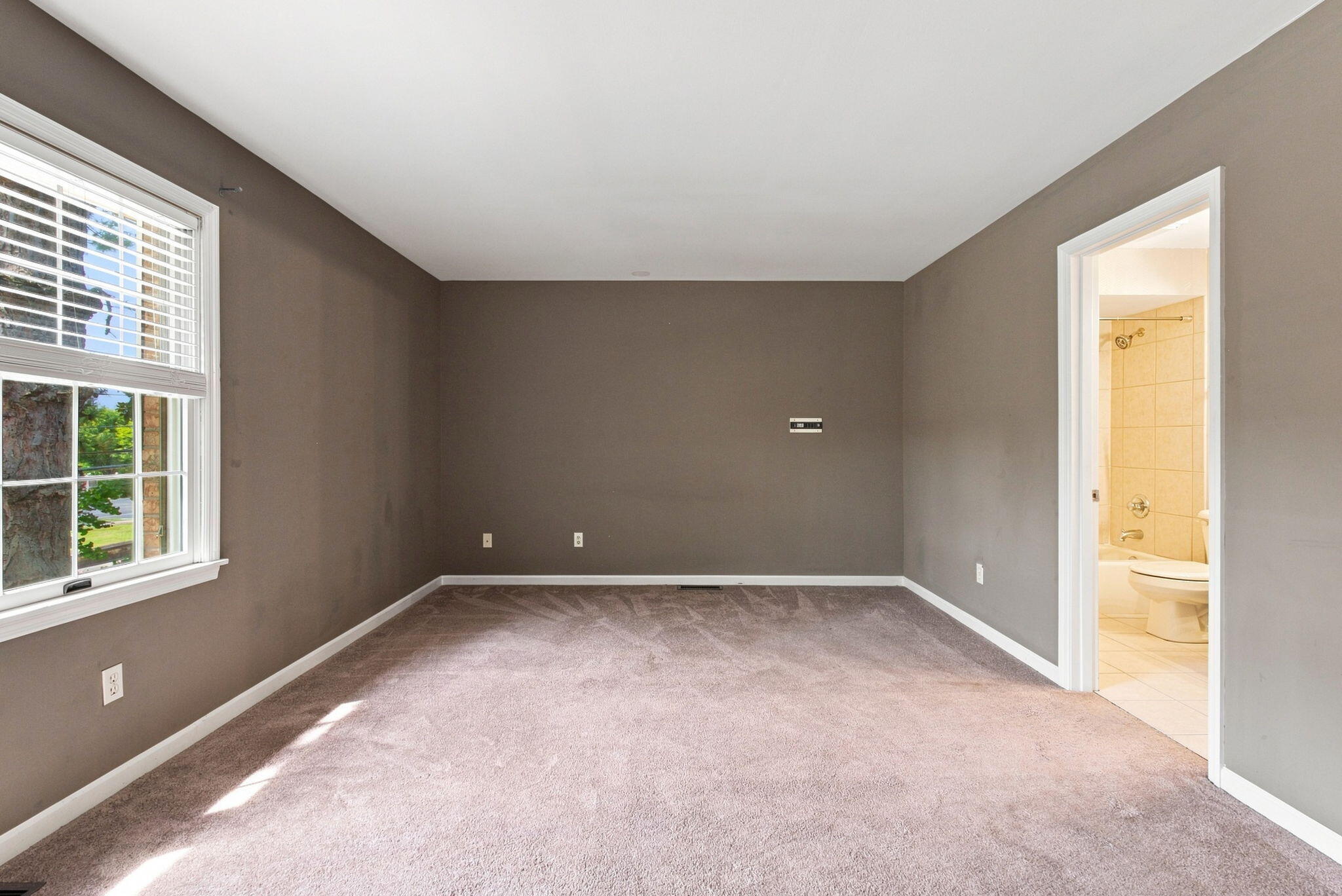 Empty carpeted bedroom with a large window and an open doorway to a tiled bathroom.