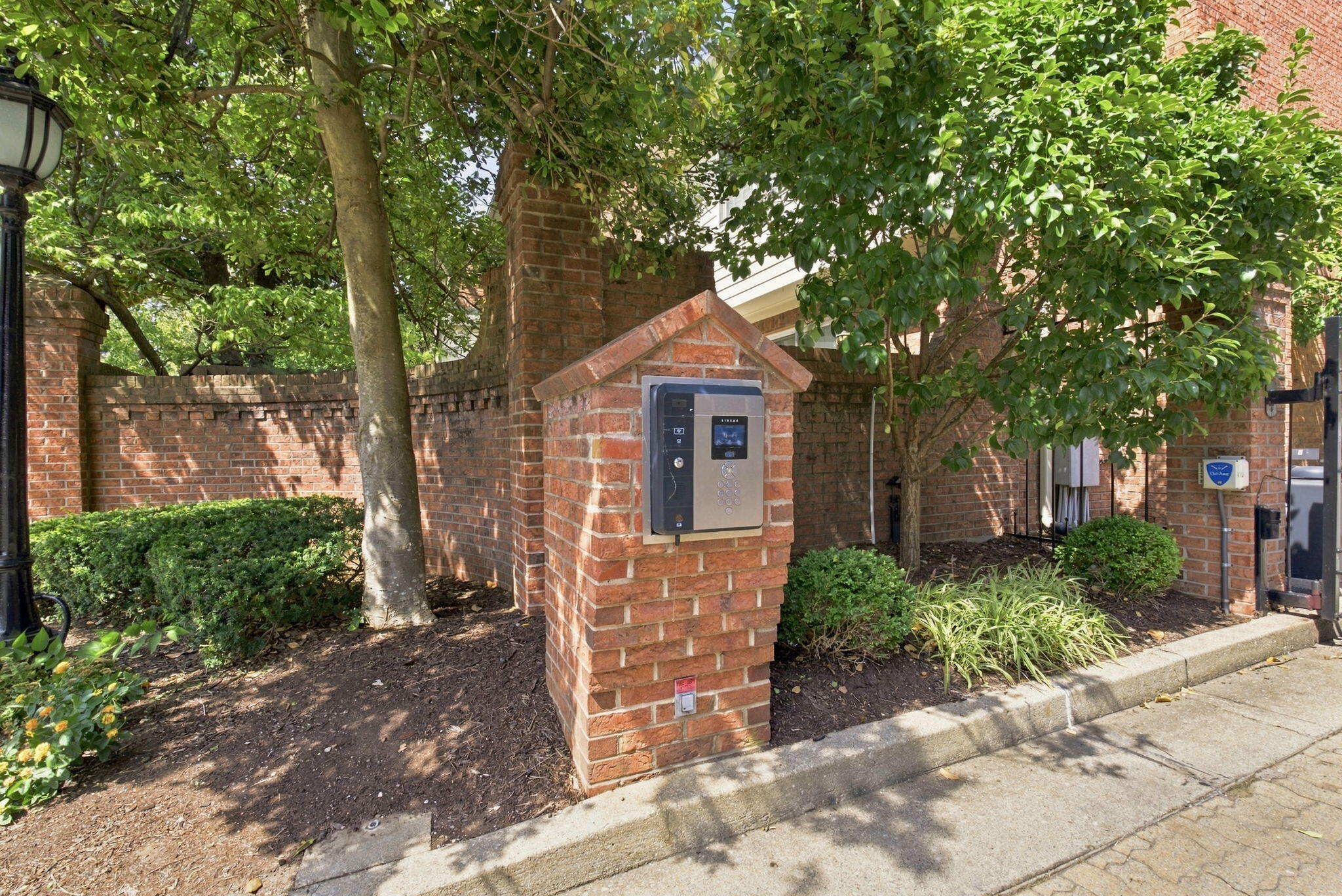 Brick building entrance with an electronic intercom pillar, mature trees, and landscaped shrubs.