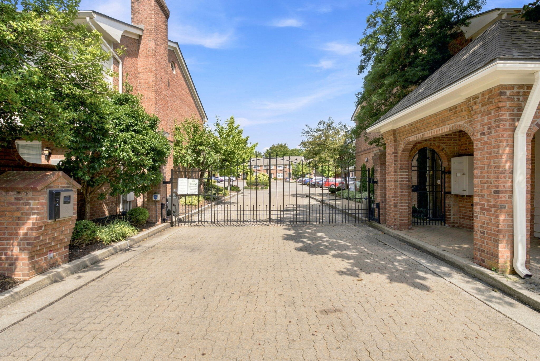 Gated residential complex entrance with brick gatehouse, black metal gate, intercom, and paved driveway.