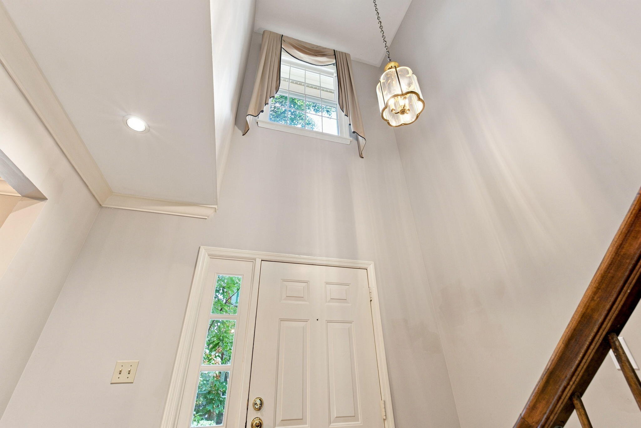 Tall entryway with a paneled front door, high curtained window, hanging light, and wooden stair railing.