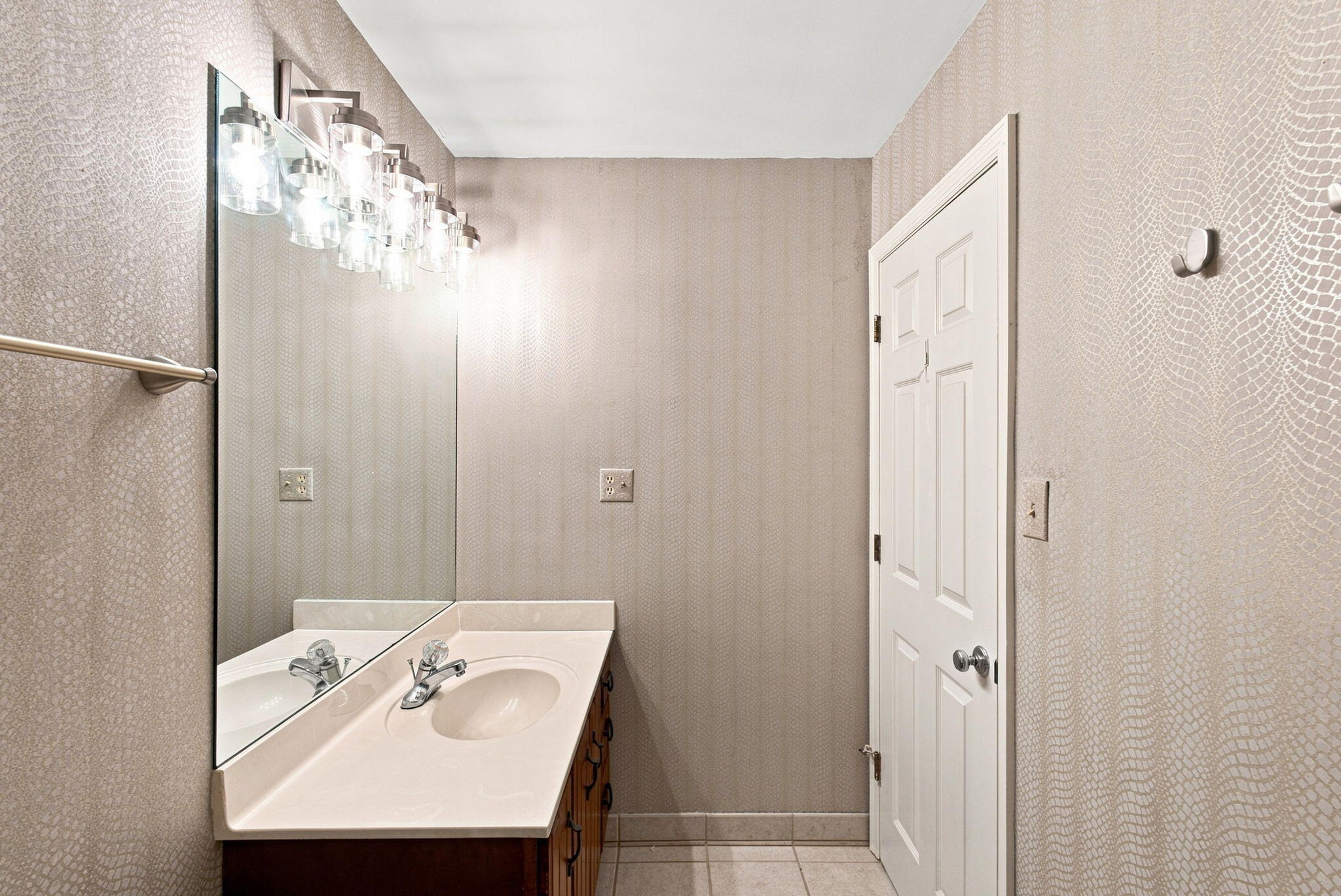 Bathroom with a white vanity, large mirror, bright multi-light fixture, and patterned beige walls.