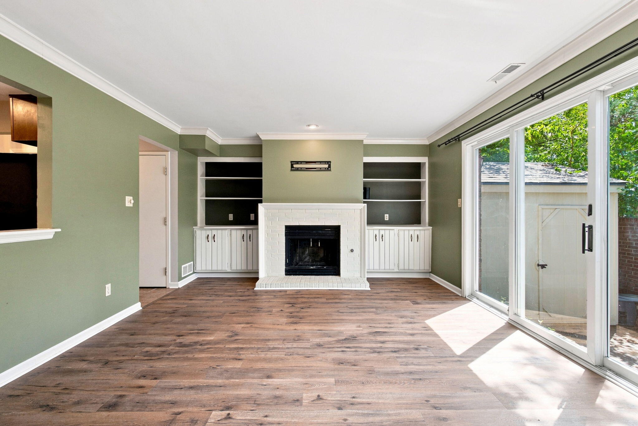 Vacant living room with a white brick fireplace, built-in shelves, wood floors, and sliding glass doors.