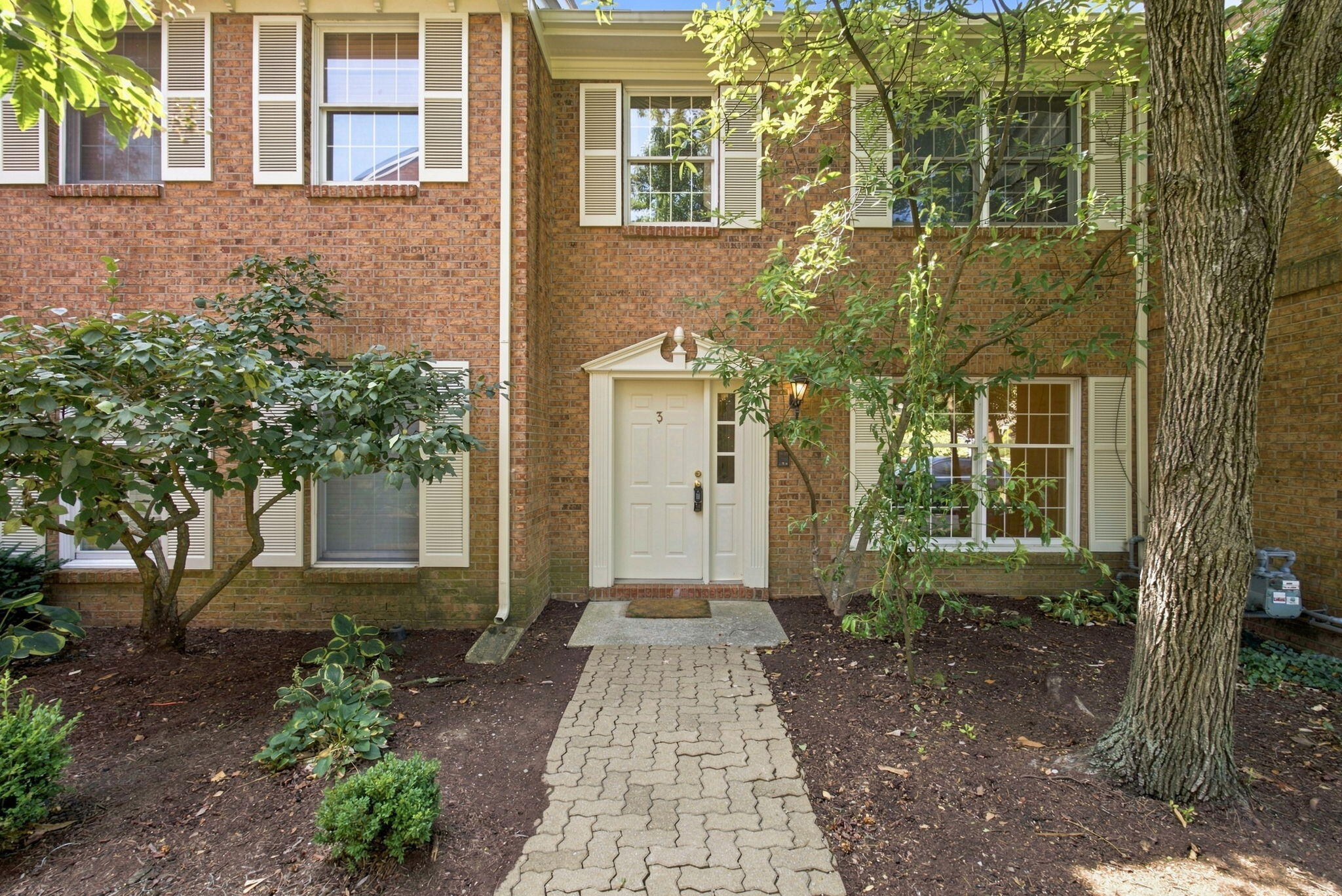 Brick home entrance with white shutters, a paved walkway, and landscaped garden beds.