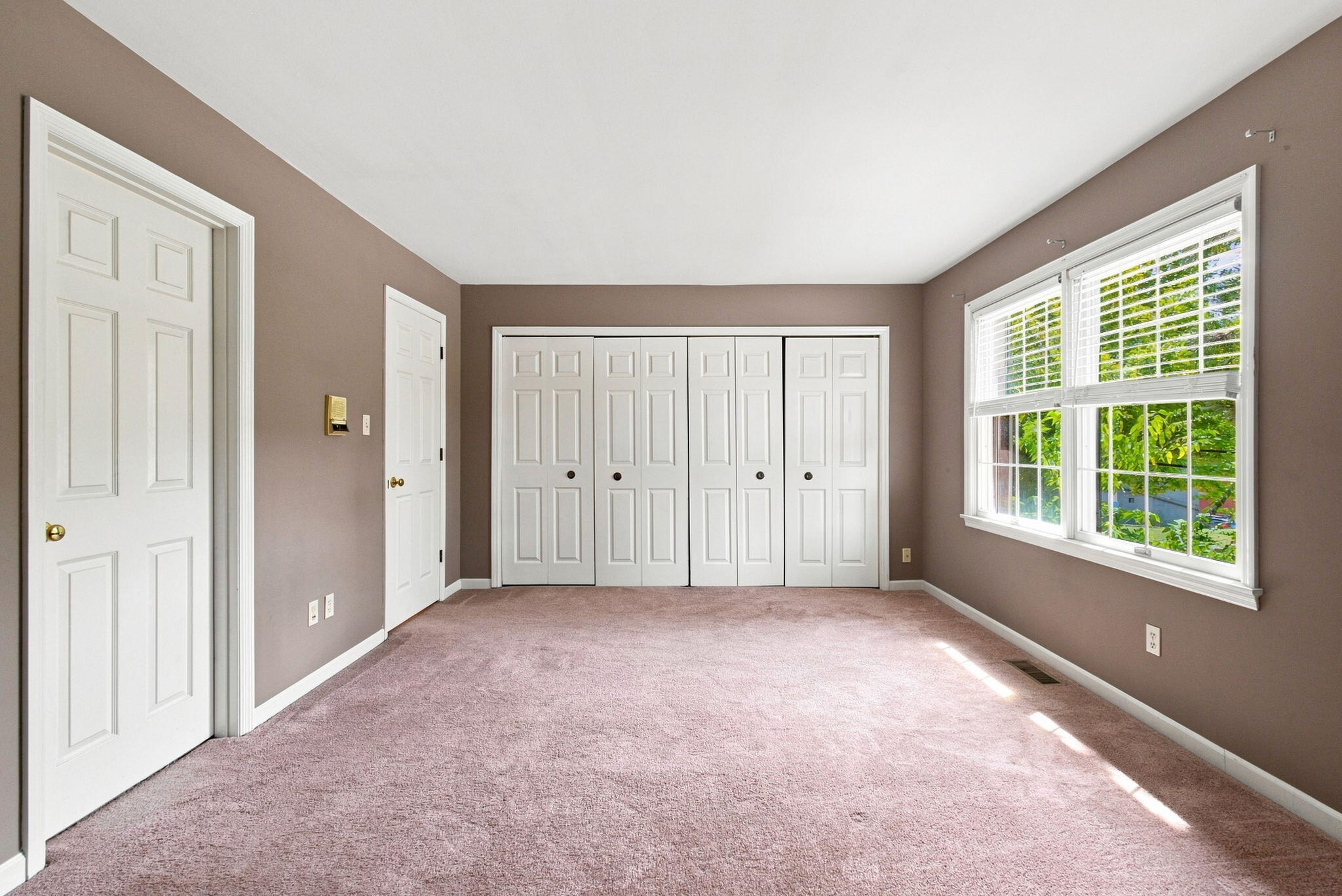 Empty bedroom with white paneled closet doors, mauve carpet, and a sunlit window with blinds.