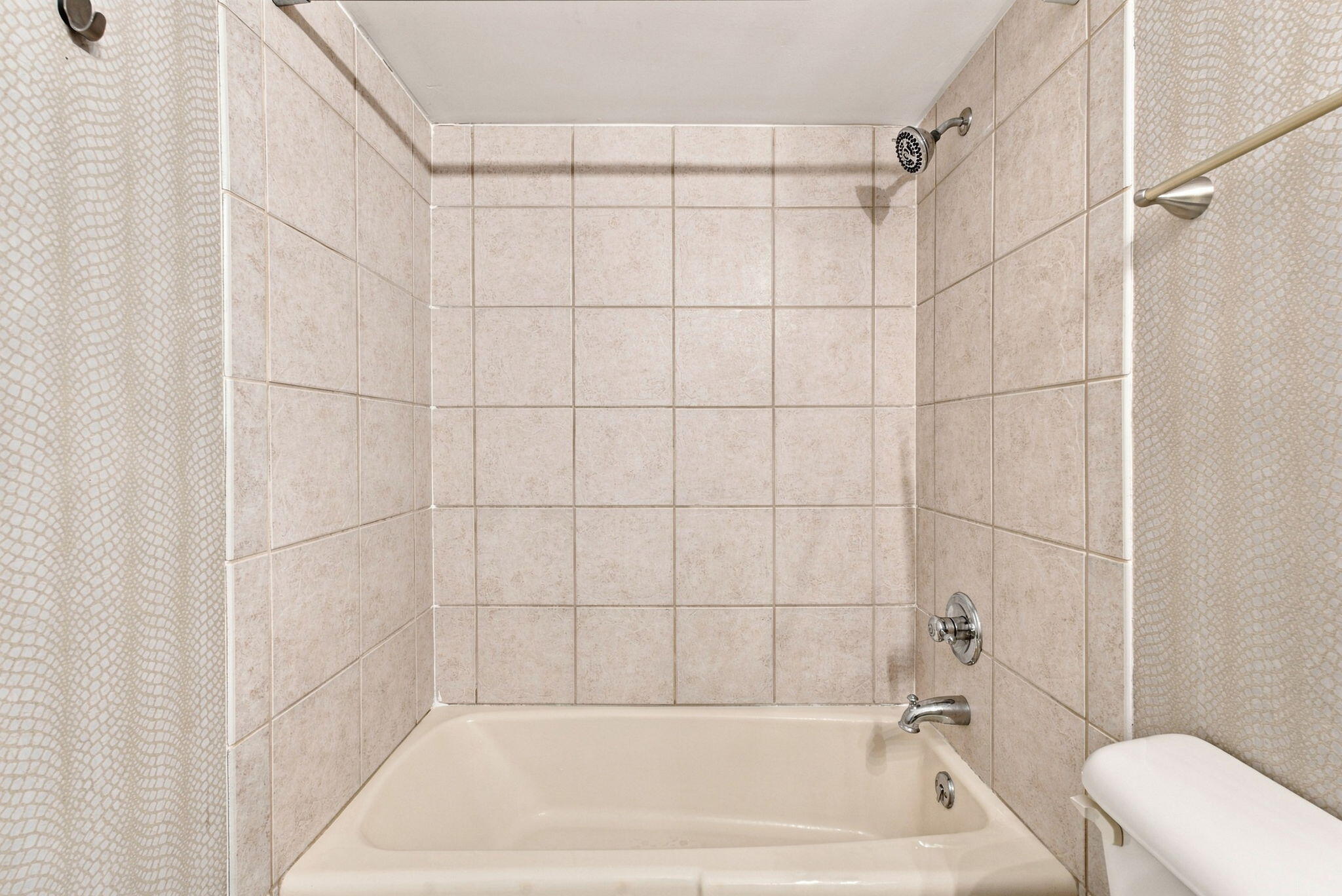 Bathroom bathtub beneath a beige tiled shower surround, with a patterned curtain and chrome fixtures.