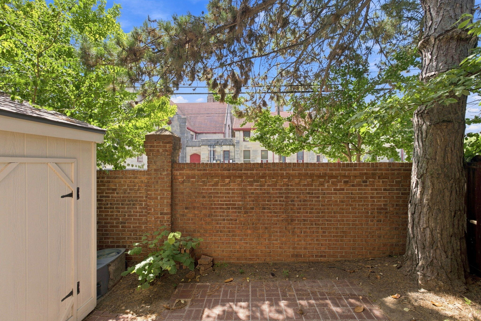 Brick-paved courtyard with a tall brick wall, white shed, and mature trees.