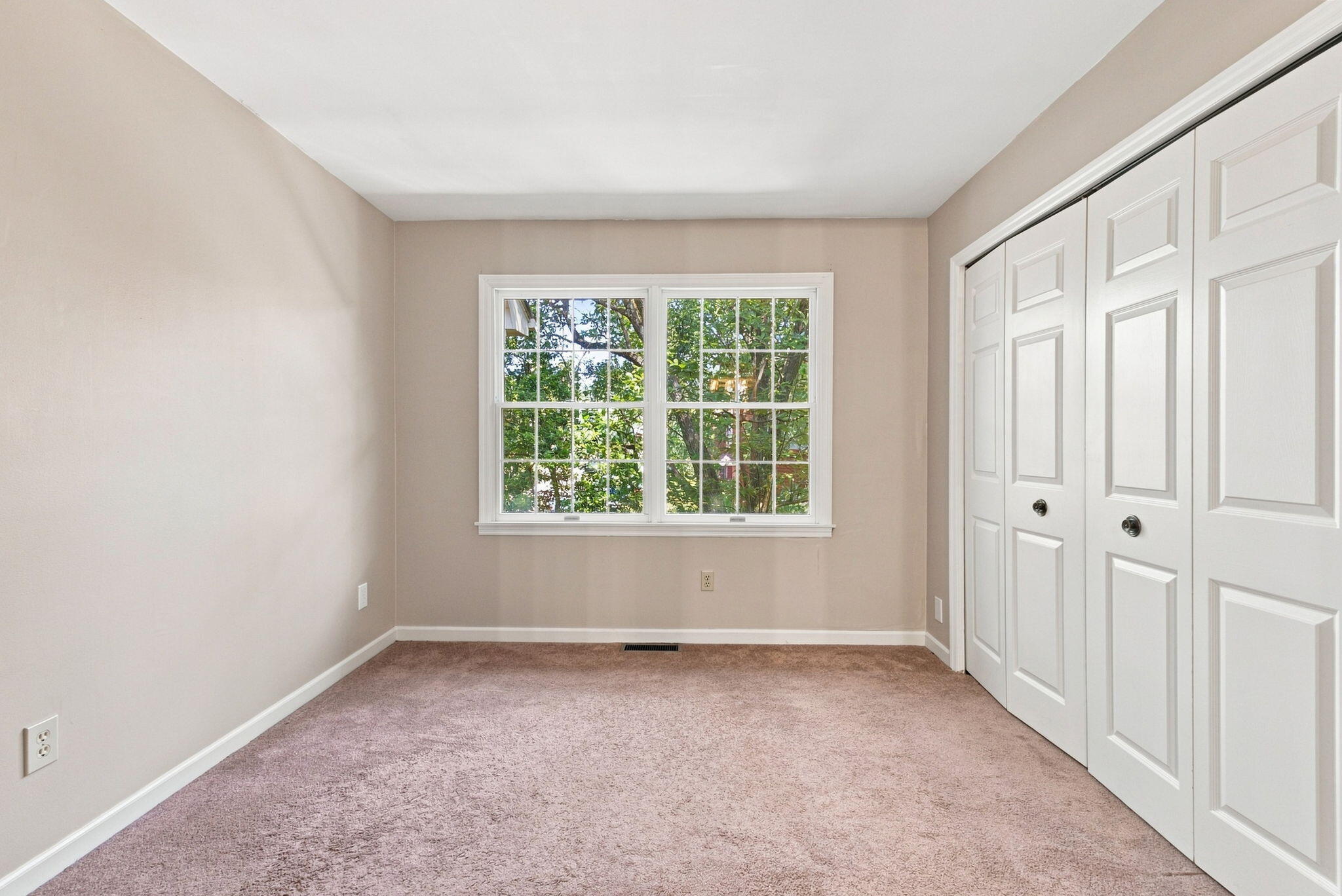 Empty bedroom with mauve carpet, leafy window view, and white paneled closet doors.