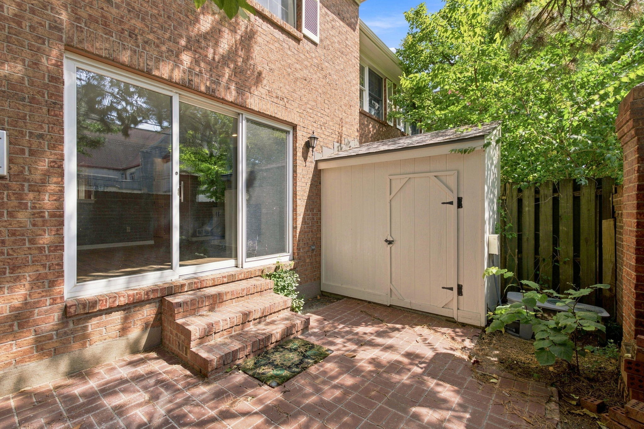 Brick patio beside a brick building, with sliding doors, brick steps, and a light-colored storage shed.