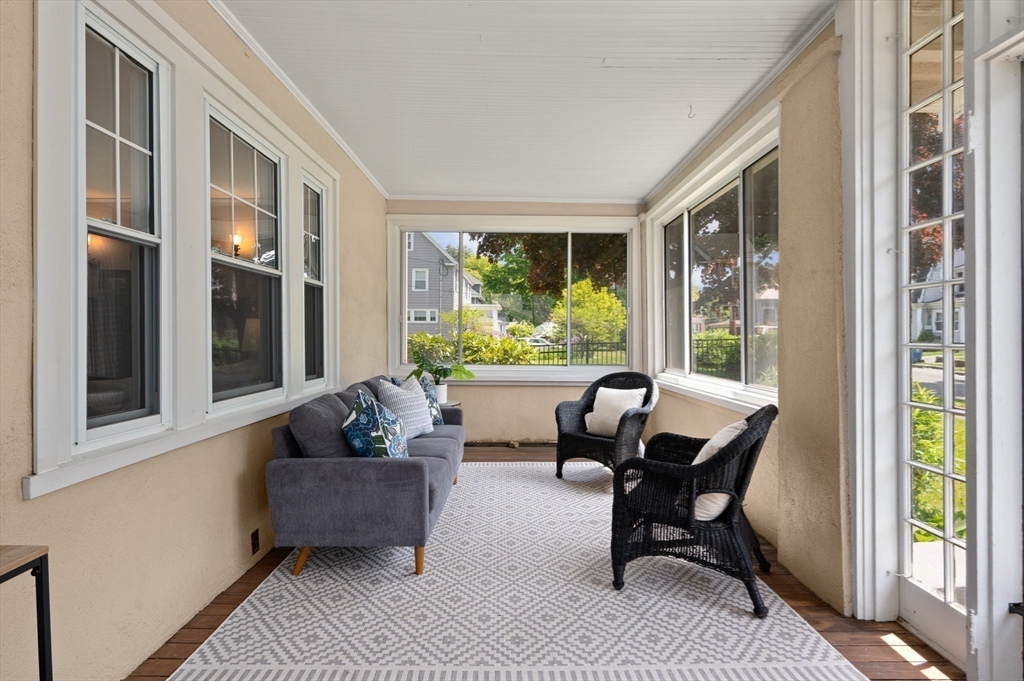 Enclosed sunroom with large windows, a gray sofa, wicker chairs, and a patterned rug.