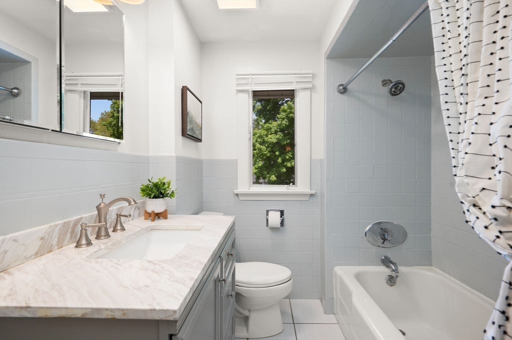 Bathroom with marble-look vanity, gray tile, and a bathtub beside a window overlooking trees.
