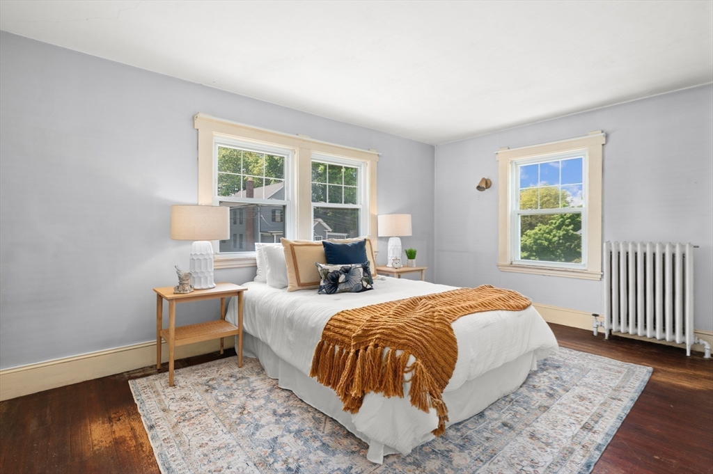 Staged bedroom with a white bed, rust-colored throw, patterned rug, and windows overlooking greenery.