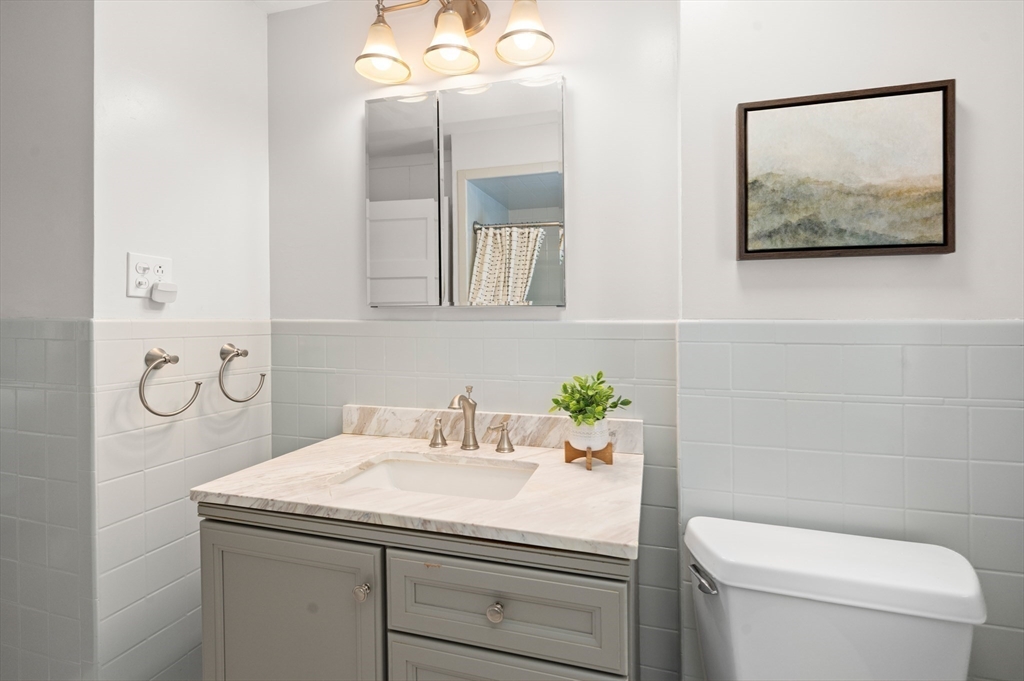 Bathroom with gray vanity, marble-look counter, mirrored cabinet, white tile, and framed landscape artwork.