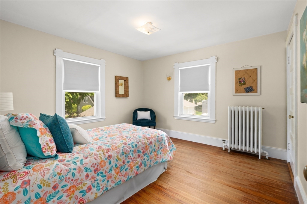 Bedroom with colorful floral bedding, hardwood floors, two shaded windows, and a white radiator.