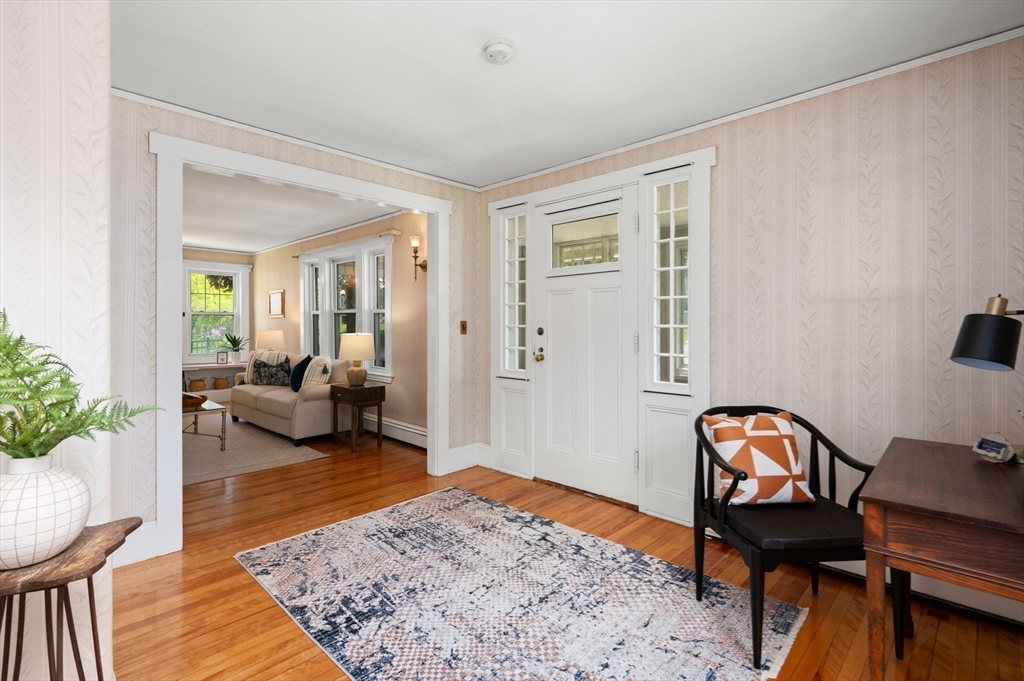 Furnished entryway with hardwood floors, a white front door, patterned wallpaper, and a view into the living room.