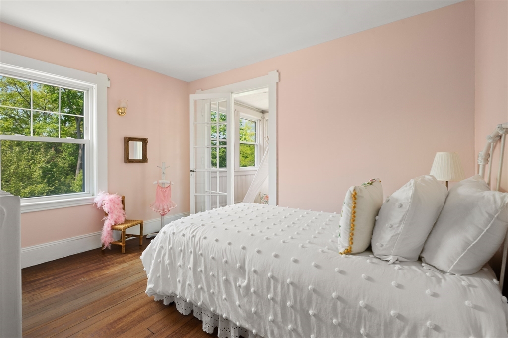 Bedroom with a white bed, pale pink walls, wood floors, and windows overlooking leafy trees.