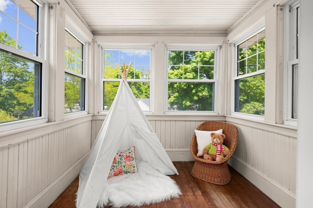 Bright sunroom with a white teepee, floral pillow, and wicker chair beside leafy windows.