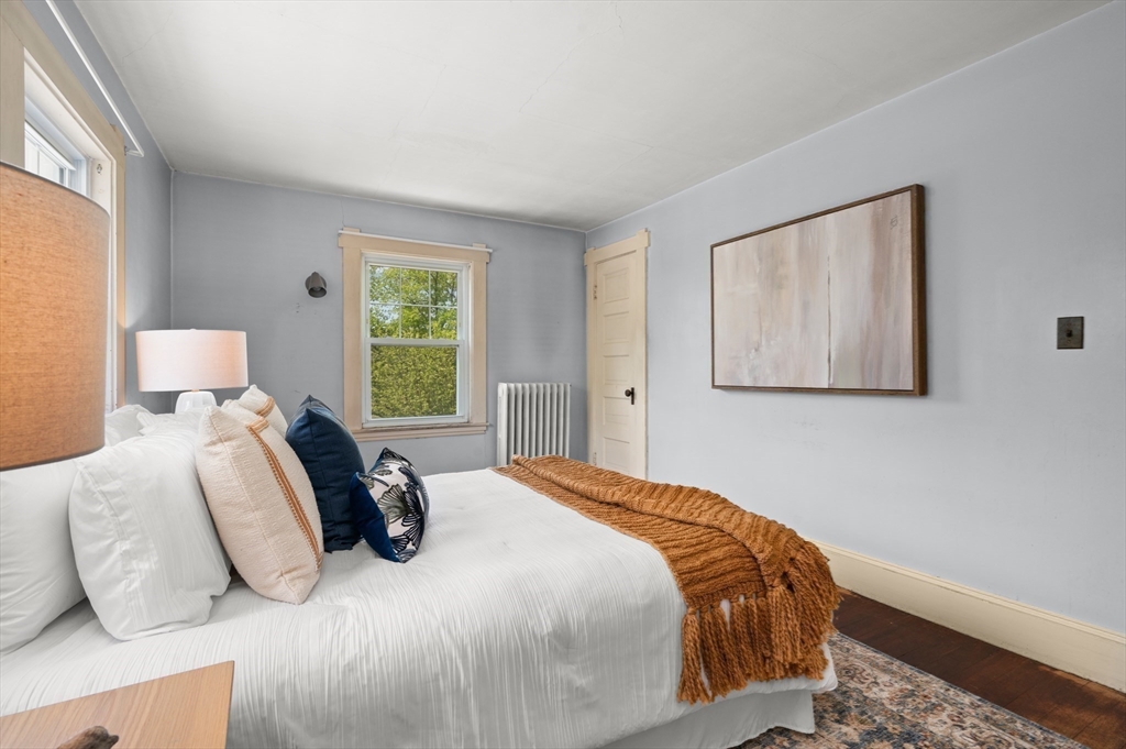 Staged bedroom with a white bed, rust-colored throw, gray walls, and a window overlooking greenery.