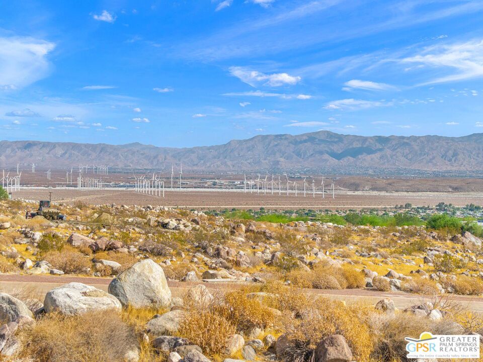 Desert valley with wind turbines, rocky scrubland, and mountains beneath a blue sky.