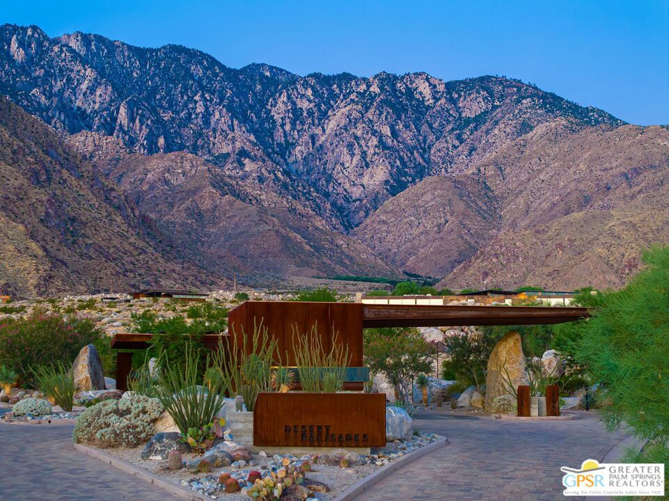 Landscaped entrance with rust-colored signs and metal structures, backed by rugged desert mountains.