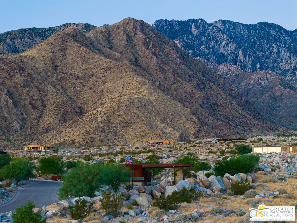 Desert property entrance beneath rugged mountains, with boulder landscaping and an American flag.
