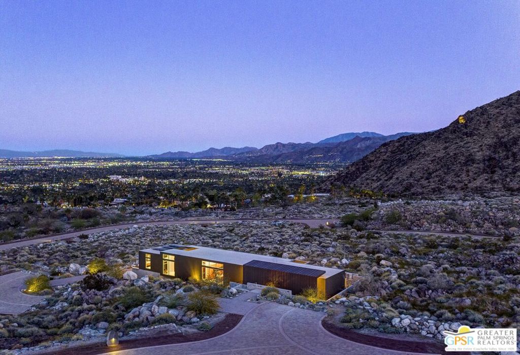 Modern desert home glowing at dusk, with Palm Springs city lights and mountains beyond.