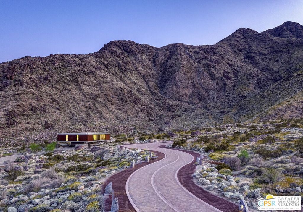 Modern desert home beside a winding road, with rugged mountains behind it at dusk.