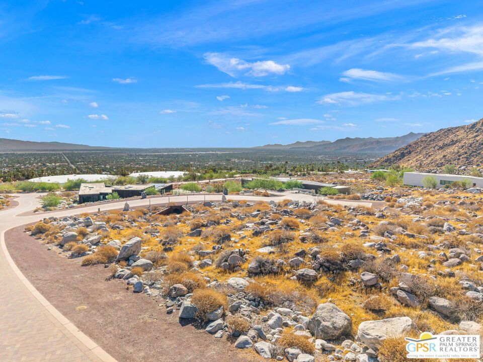 Rocky desert hillside overlooks the Palm Springs valley, with modern homes and distant mountains.