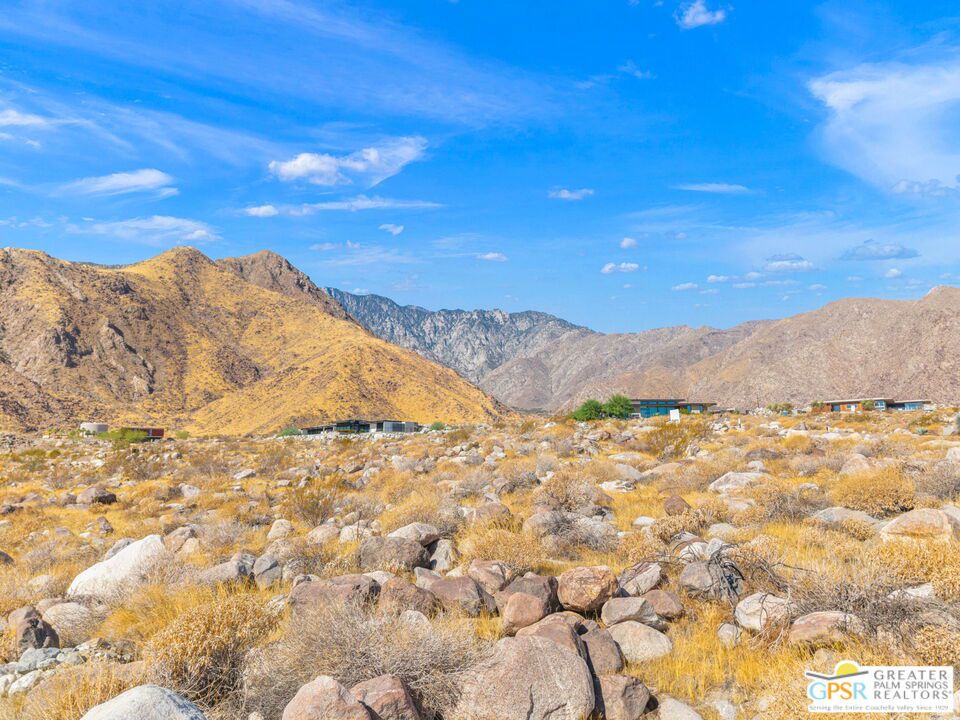 Desert boulders and scrub in the foreground, with mountain ranges and homes beyond.