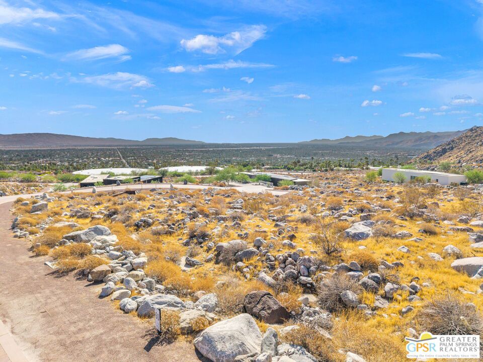 Vacant rocky desert lot with golden scrub, an access road, and distant valley and mountain views.