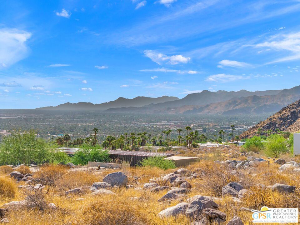 Desert property view over Palm Springs, with rocky foreground, palm trees, valley, and mountains.