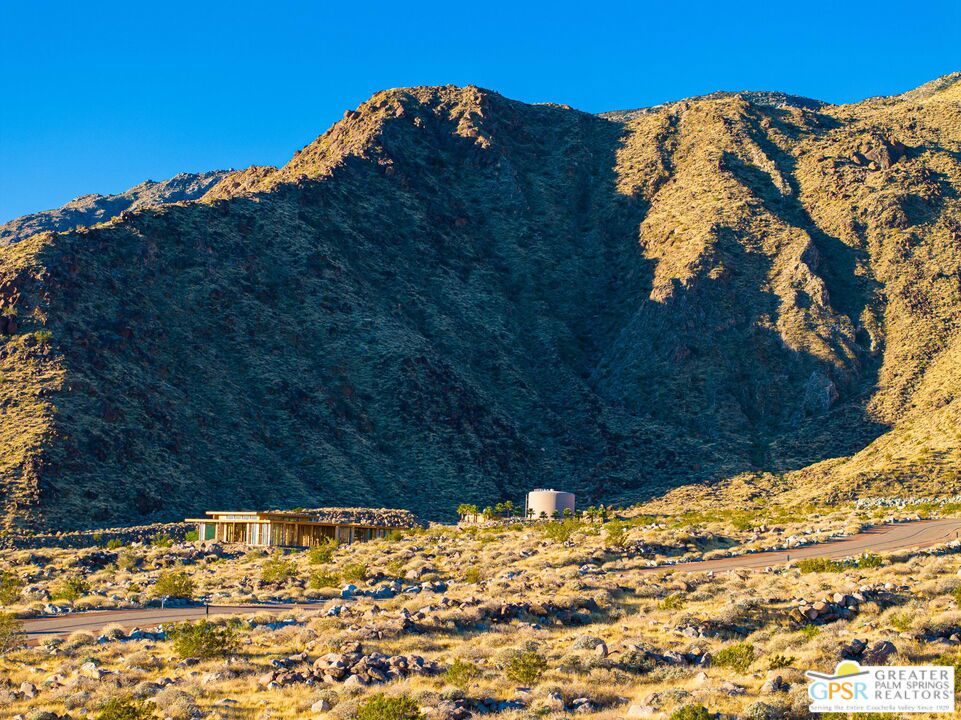 Desert mountains rise behind scrubland, a road, and a small low structure in warm sunlight.