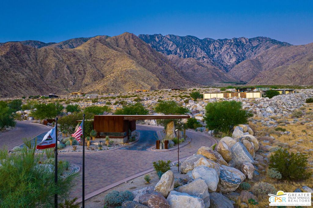 Desert community entrance with California and U.S. flags, rocky landscaping, and rugged mountains.