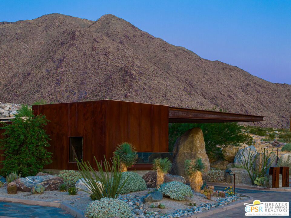 Modern rust-colored home with a cantilevered roof, desert landscaping, and mountain backdrop.