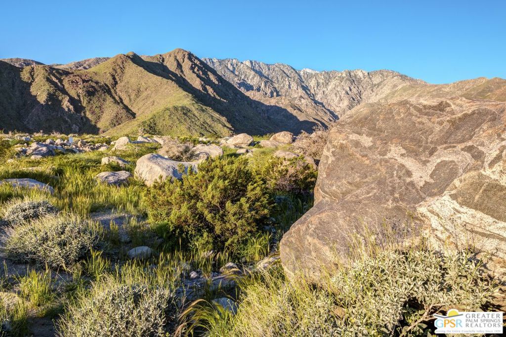 Rocky desert scrub and boulders below rugged mountains in warm sunlight.