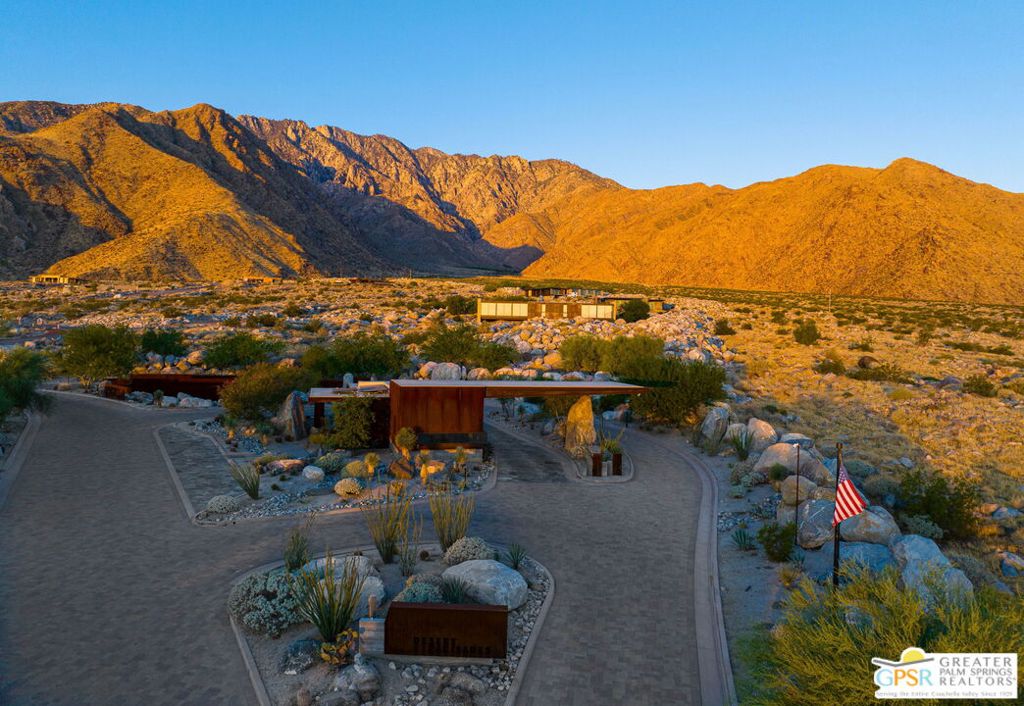 Desert property entrance with winding drive, boulders, American flag, and golden-lit mountains.