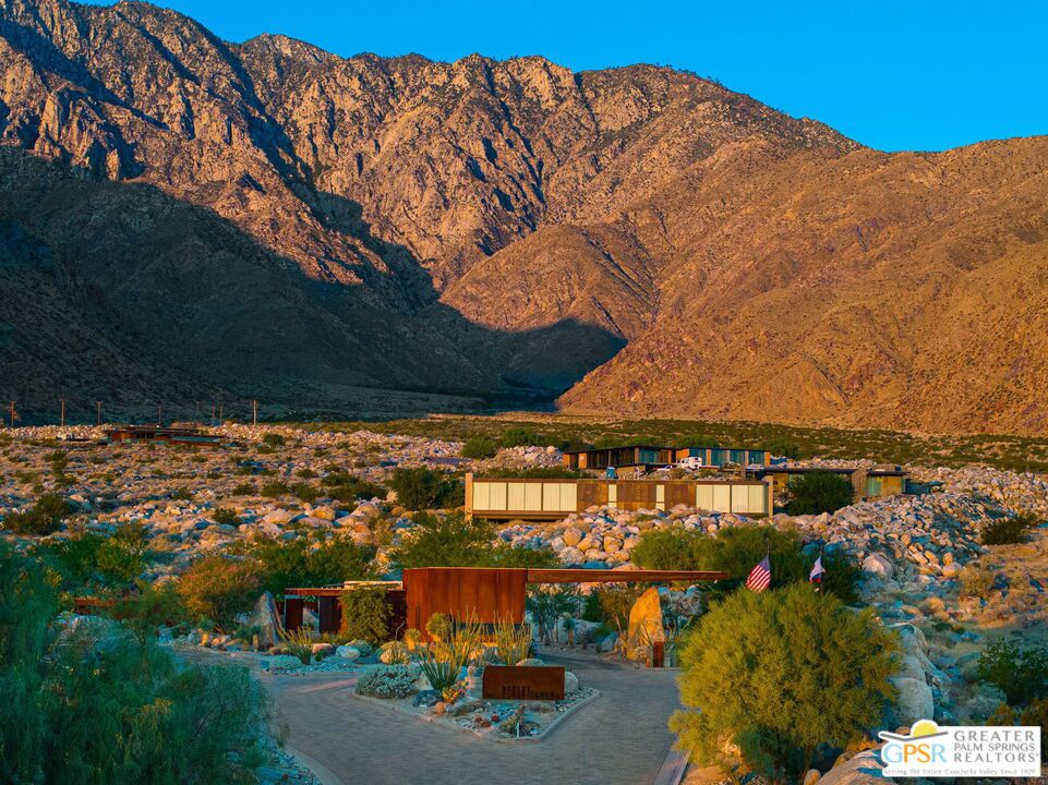 Desert home beneath sunlit mountains, with rocky landscaping and a long modern facade.
