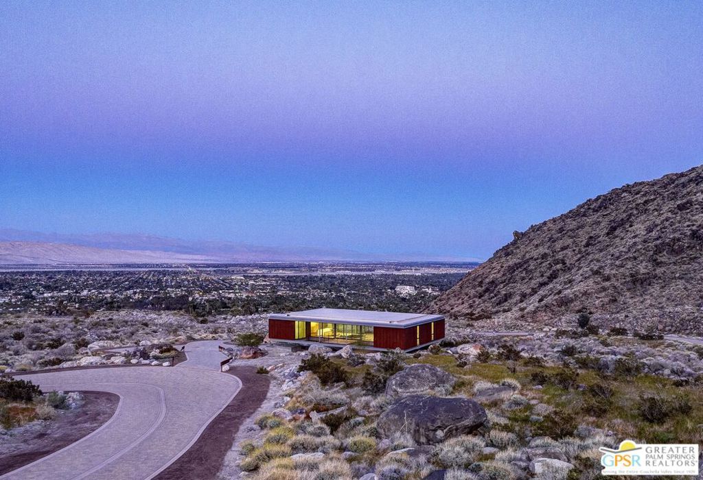 Modern desert home with red façade, curved driveway, mountain slope, and Palm Springs valley view.