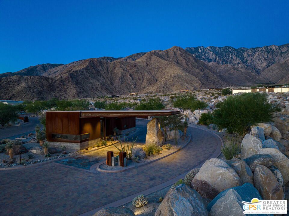 Modern desert home with a paved drive, rocky landscaping, and mountains behind it at dusk.